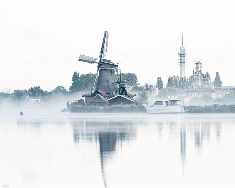 Historic windmill reflected in calm water and surrounded by dense morning mist in a high-key photograph at Zaanse Schans, the Netherlands.