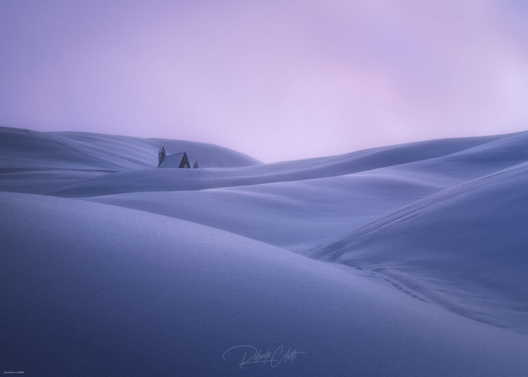 Small chapel surrounded by wind-shaped snowdrifts at Giau Pass during winter in the Italian Dolomites.
