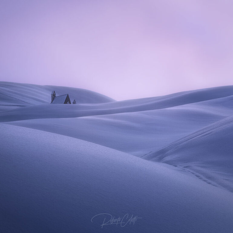Small chapel surrounded by wind-shaped snowdrifts at Giau Pass during winter in the Italian Dolomites.