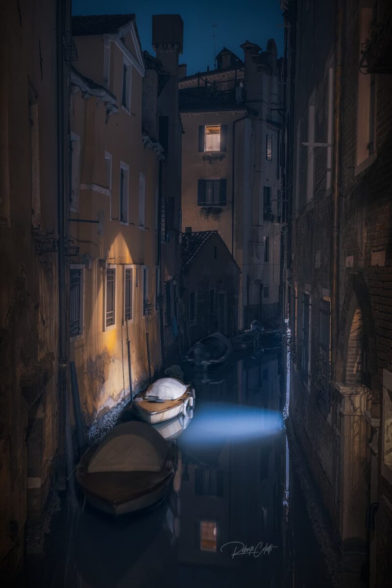 An illuminated boat moored along a narrow Venetian canal at night, surrounded by warm lights and historic architecture in Venice, Italy.