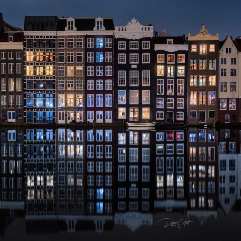 Historic canal houses illuminated at night along Damrak in Amsterdam, the Netherlands, with their lights reflecting across the water.