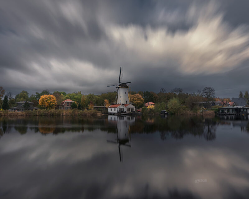 Traditional Dutch windmill reflected in calm water beneath dramatic storm clouds in the Netherlands.