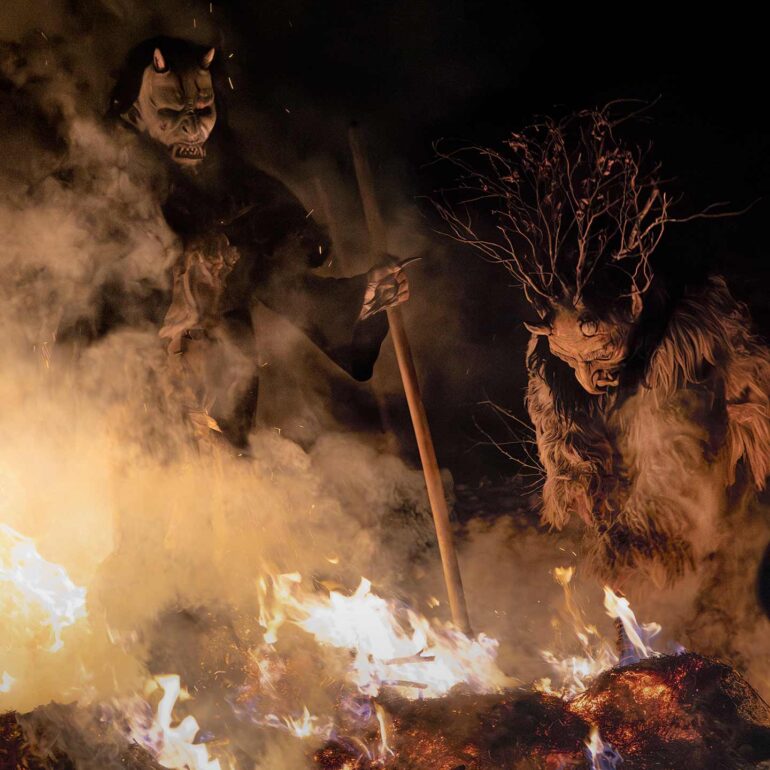 Traditional Krampus wearing a hand-carved wooden mask surrounded by fire during a winter night parade in Italy.