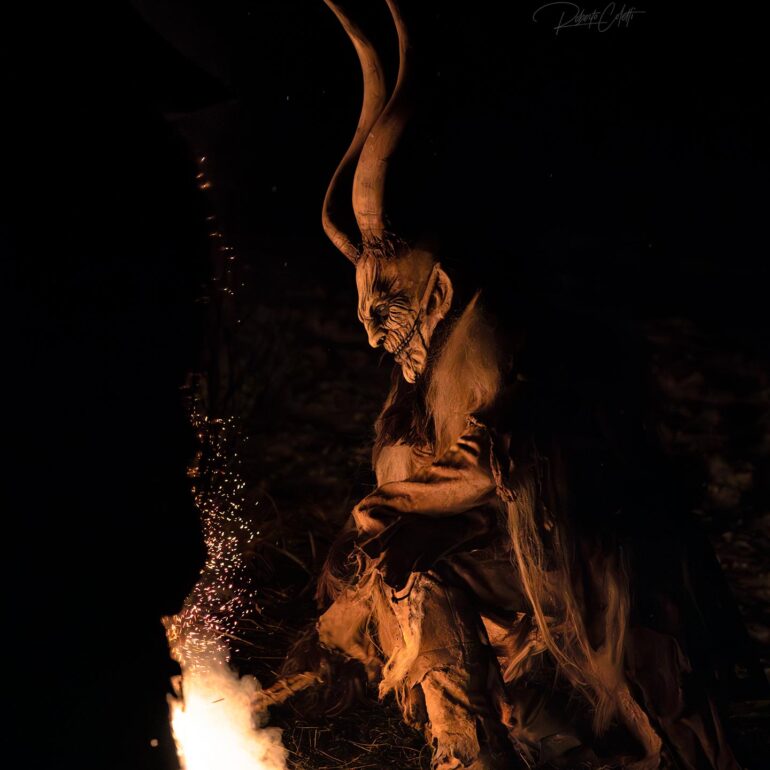 Krampus wearing large horns and a traditional wooden mask during a night celebration in the Dolomites, Italy.