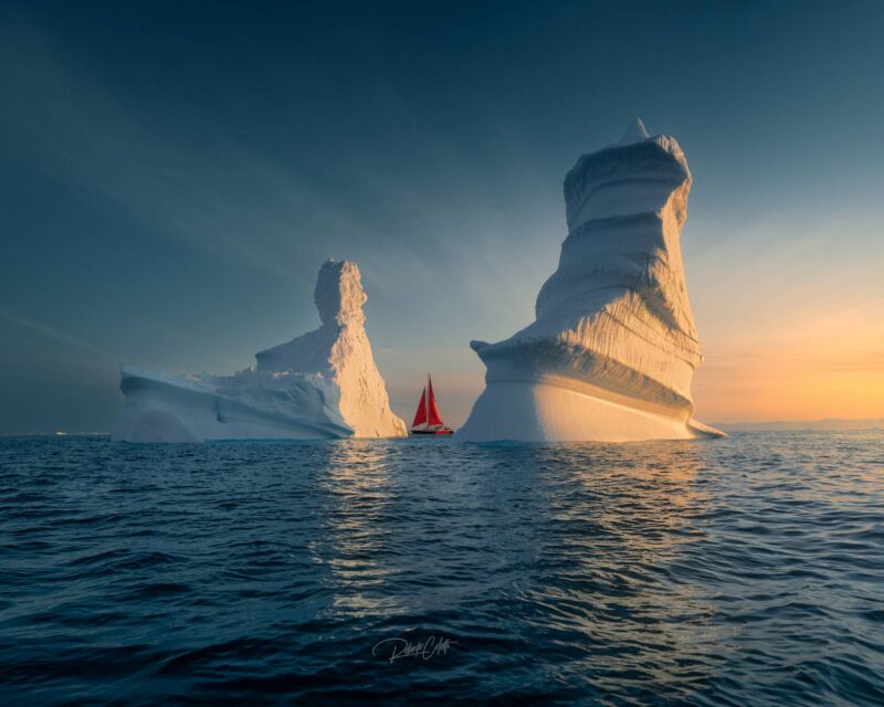 Traditional sailing ship with striking red sails navigating between two giant icebergs during summer in the Arctic waters of Greenland.