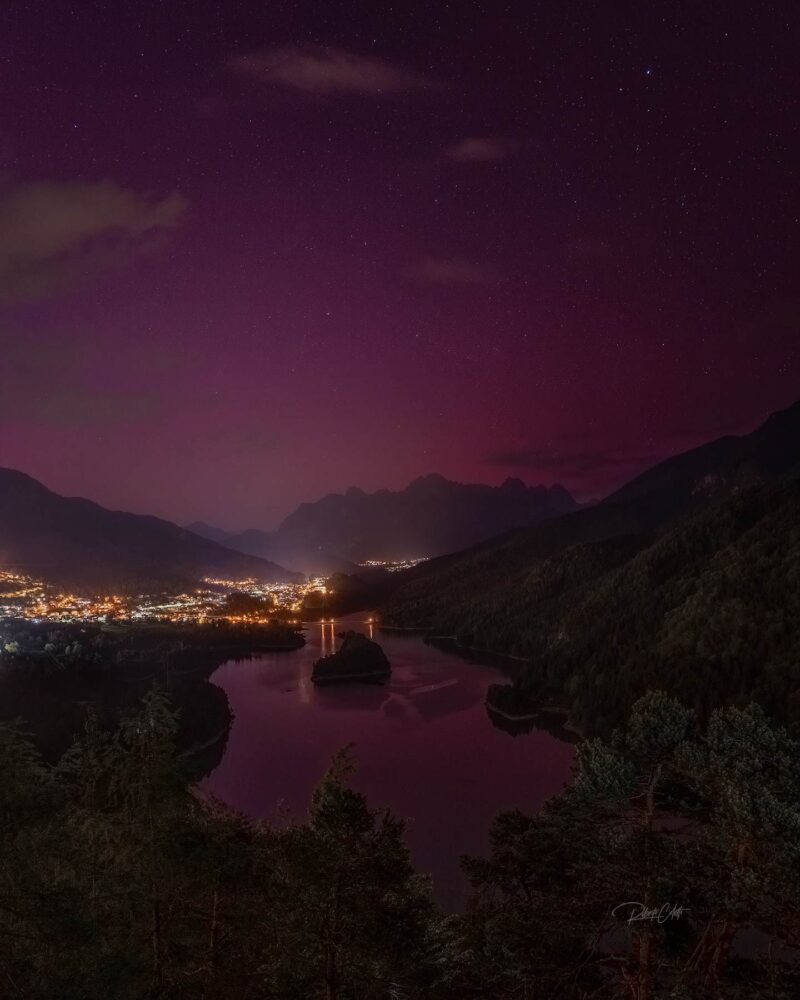 Aurora Borealis illuminating Lake Centro Cadore in the Italian Dolomites, with colorful reflections on the water during a clear night.