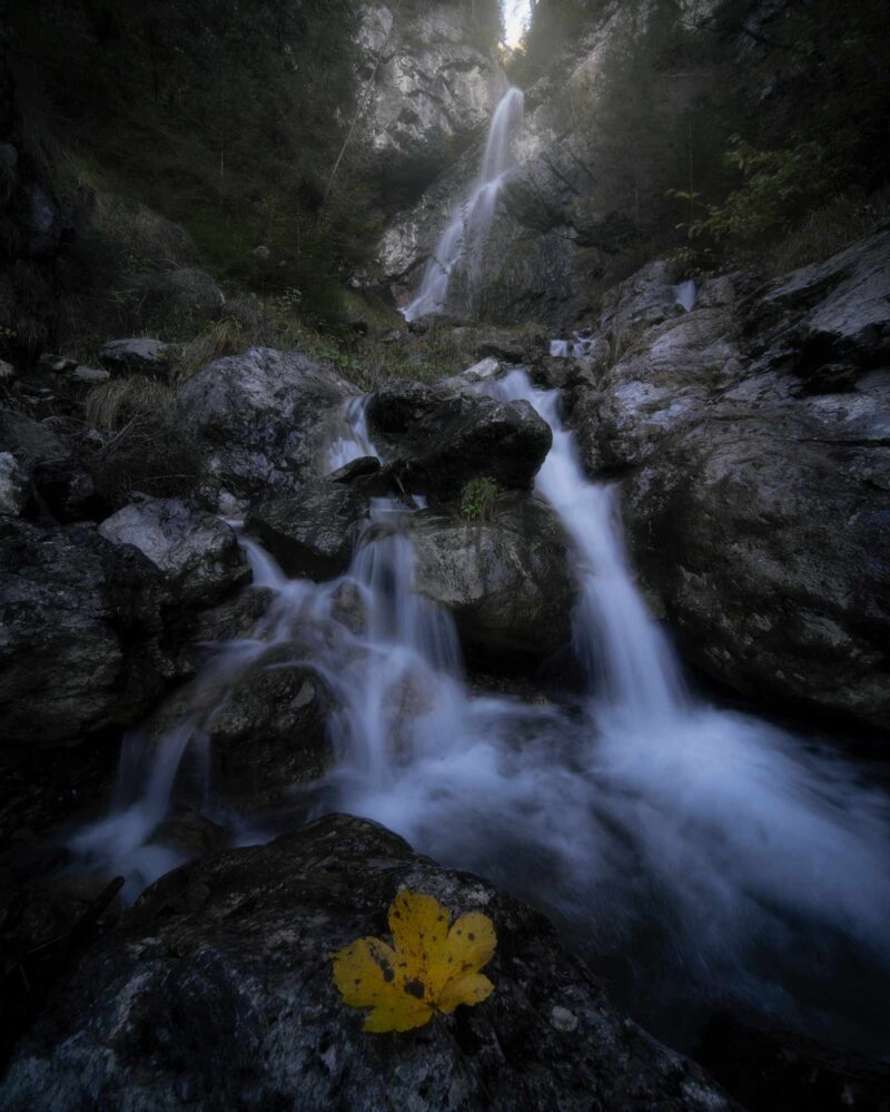 Hidden waterfall in Nebbiù di Cadore surrounded by colorful autumn forest in the Italian Dolomites, captured during the soft light of sunrise.