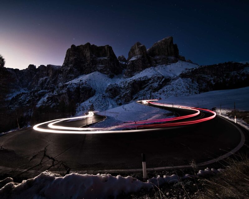 Long-exposure sunrise at Passo Gardena in the Italian Dolomites, with colorful vehicle light trails winding through the mountain road.