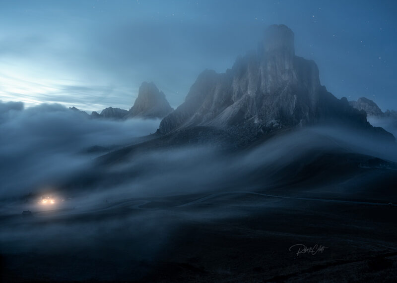 Long-exposure photograph of Passo Giau in the Italian Dolomites during the blue hour, with drifting fog and vehicle light trails winding through the mountain pass.