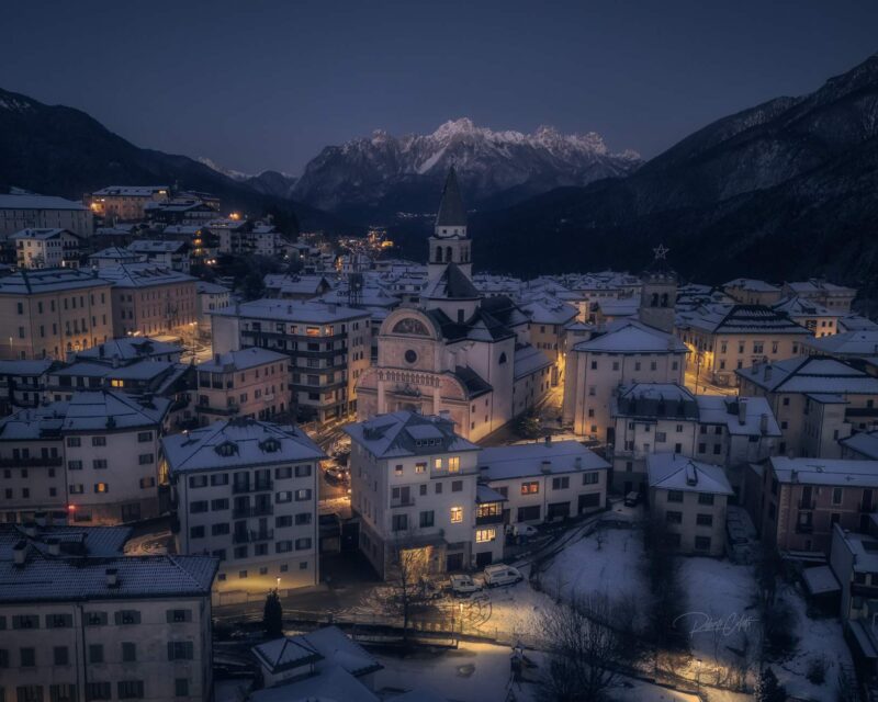 The historic center of Pieve di Cadore during the blue hour, with Monte Tudaio and the snow-covered Dolomites rising behind on a freezing winter evening in Italy.