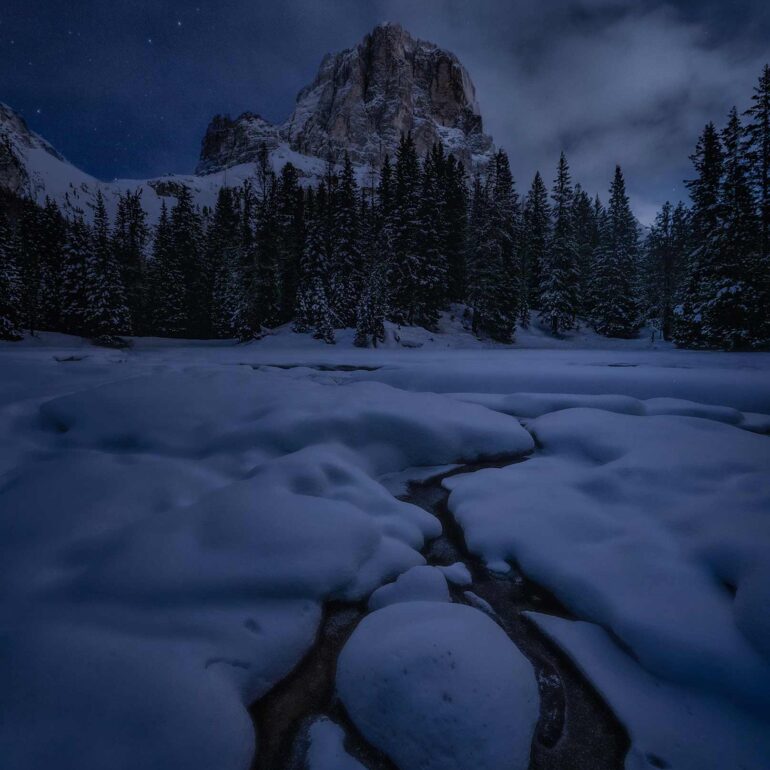 Night view of the Tofana de Rozes reflected in the snow-covered pond of Bai de Dones in the Italian Dolomites during a peaceful winter evening.