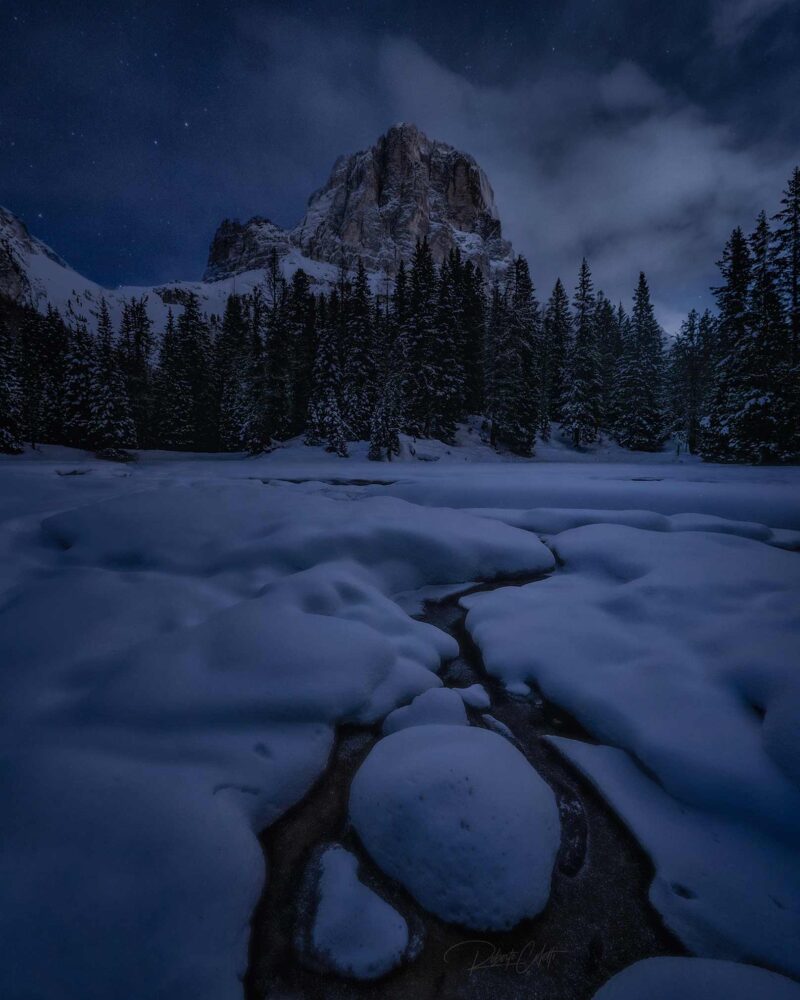 Night view of the Tofana de Rozes reflected in the snow-covered pond of Bai de Dones in the Italian Dolomites during a peaceful winter evening.
