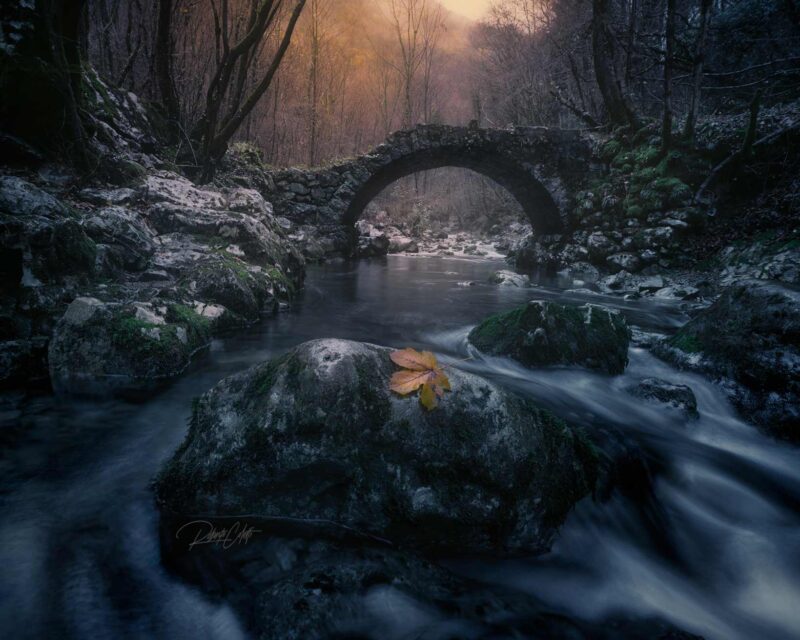 Ancient Roman bridge hidden in the forests of Friuli Venezia Giulia, Italy, crossing a fast-flowing river during an autumn sunset.