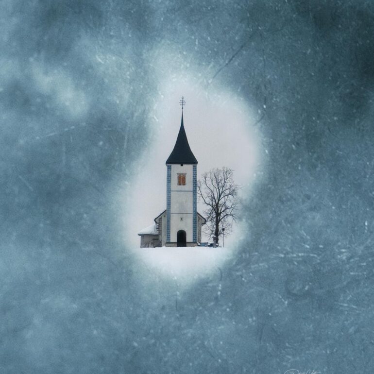 The iconic church of Jamnik in Slovenia framed through a natural opening in the snow during a peaceful winter day.