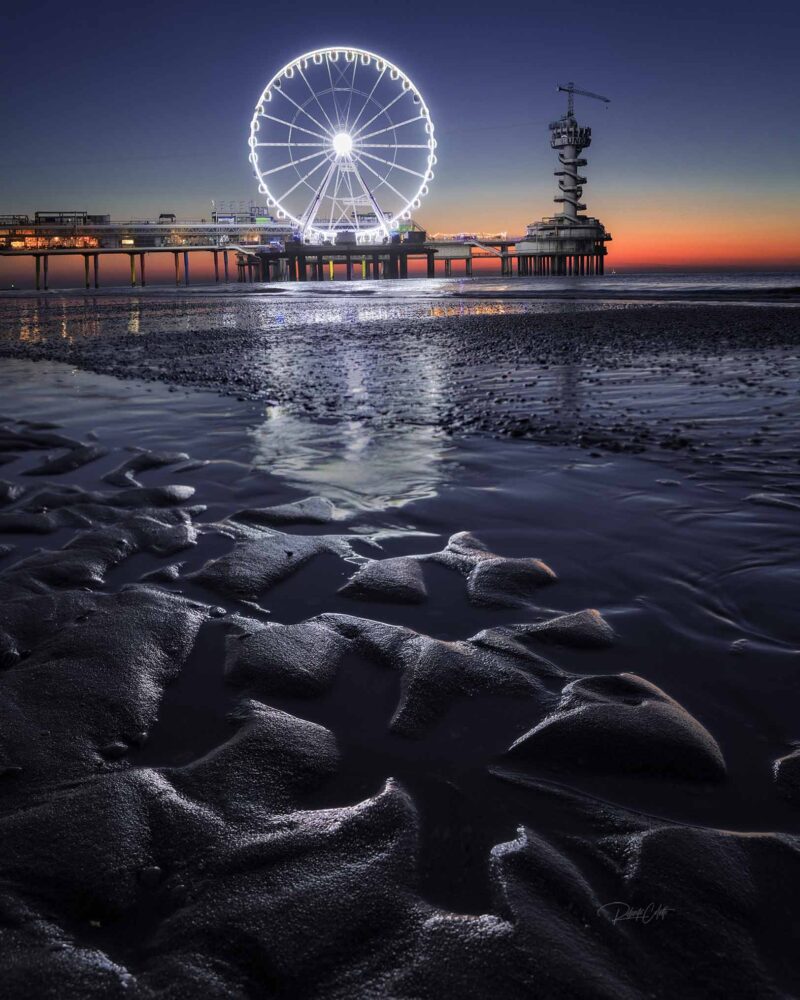 Scheveningen Pier at sunset during low tide on a summer evening, with the sandy beach exposed along the North Sea coast in the Netherlands.