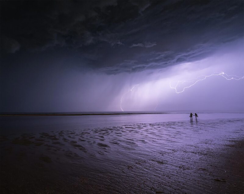 A couple holding hands while watching lightning over the beach in Noordwijk, the Netherlands, during a dramatic summer thunderstorm at sunset.